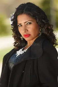 Woman with dark curly hair, red lipstick, and black coat wearing a white lace necklace.