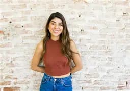Smiling woman with long brown hair wearing a rust-colored sleeveless top against a white brick wall.