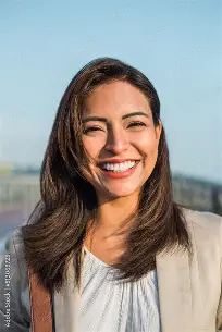 Smiling woman with long brown hair wearing a light blazer against a clear blue sky.