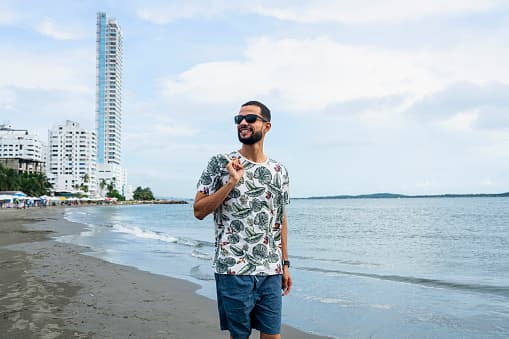 Smiling man in sunglasses and floral shirt walking on a beach near tall city buildings.